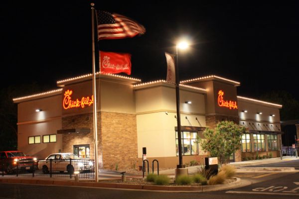 A brightly lit Chick-fil-A restaurant at night, with the American flag and Chick-fil-A flag waving in front. The building is accented with warm lighting and stone detail.