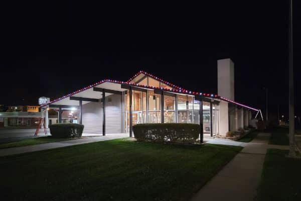 Modern commercial building at night, decorated with red and blue Christmas lights along the roofline. Bushes in front, dark sky above.
