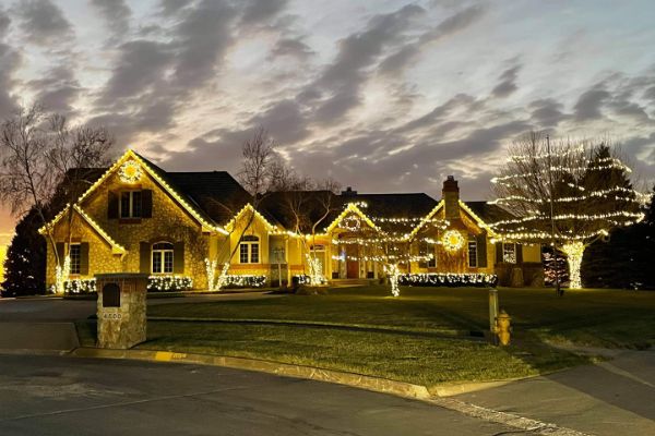 Large home with warm white lights along the rooflines, windows, and trees at dusk.