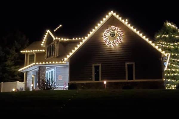 House with warm white roofline lights, a wreath, and an illuminated evergreen tree.