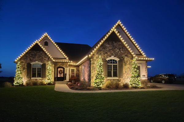 Stone and stucco home with warm white roofline lights and illuminated evergreen trees by the entrance.