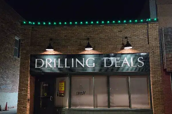 The storefront of "Drilling Deals" is illuminated at night, with green lights above the brick facade. Three spotlights shine on the sign above the windows.