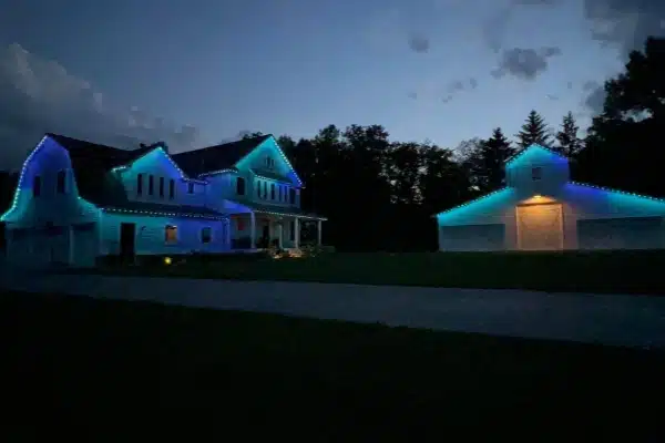A two-story house and barn are outlined with blue and white holiday lights against a twilight sky. The buildings sit on a large lawn, creating a festive and inviting scene.