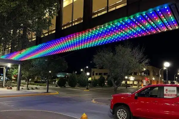 A vibrant canopy of rainbow-colored lights illuminates the entrance of a building at night. A red pickup truck is parked nearby, adding a pop of color to the urban scene.