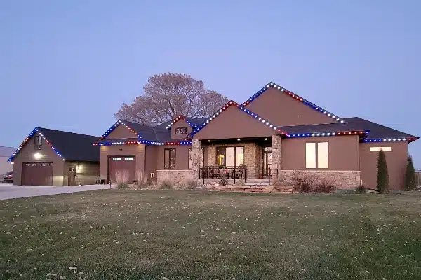 Suburban home at dusk adorned with patriotic red, white, and blue lights along the roofline. Stone accents highlight the house's exterior, and a well-manicured lawn completes the scene.