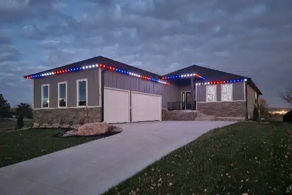 A modern home is adorned with red, white, and blue lights along the roofline against a cloudy evening sky. A concrete driveway leads to a two-car garage, with well-maintained lawn and stone accents on the facade.