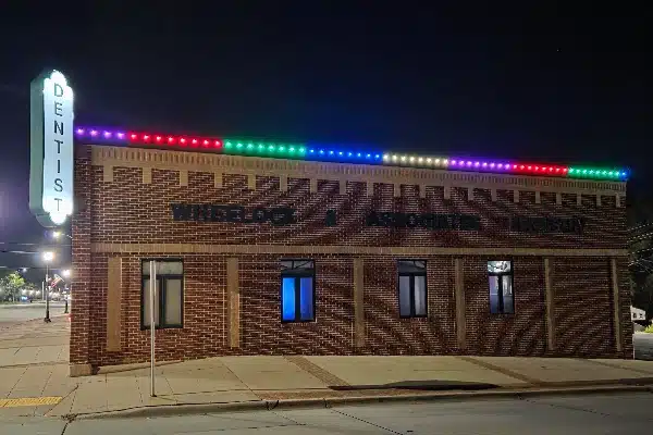 Night view of Wheelock & Jasso Dentistry building, lit with colorful lights. A tall "Dentist" sign glows beside the brick building.
