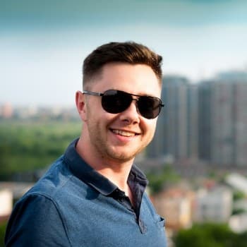 Smiling man in sunglasses and blue polo shirt stands on a rooftop, with a cityscape and green parkland in the background.