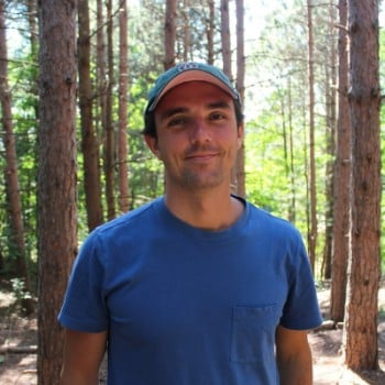 A smiling man in a blue t-shirt and green cap stands amidst tall pine trees. The sun filters through the trees, creating a peaceful, natural scene.