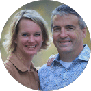 Smiling couple standing close outdoors; woman with short blonde hair in tan jacket, man in patterned light-blue shirt with arm around her.