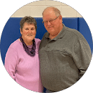 Two smiling adults side-by-side against a blue wall; left in pink sweater and scarf, right in gray sweater and glasses.